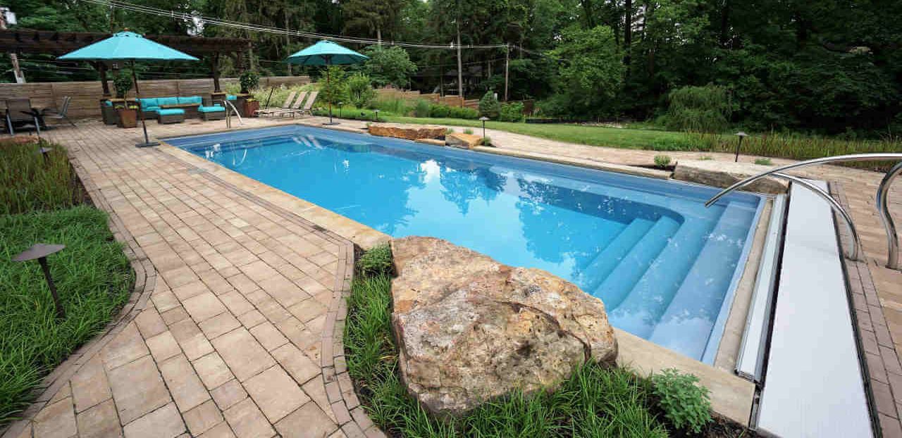 Eye-level length perspective of a rectangular blue fiberglass pool featuring entry steps at both ends, an integrated automatic safety cover housing, curved stainless steel handrails, interlocking stone paver patio decking, and large decorative boulders.