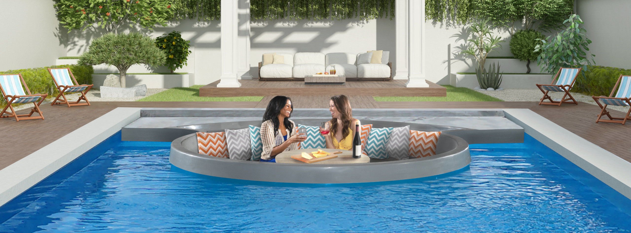 Eye-level view of two women relaxing with drinks and a cheese platter inside a round sunken living room lounge built into a rectangular blue fiberglass swimming pool, set against a modern outdoor patio with a pergola and lawn.
