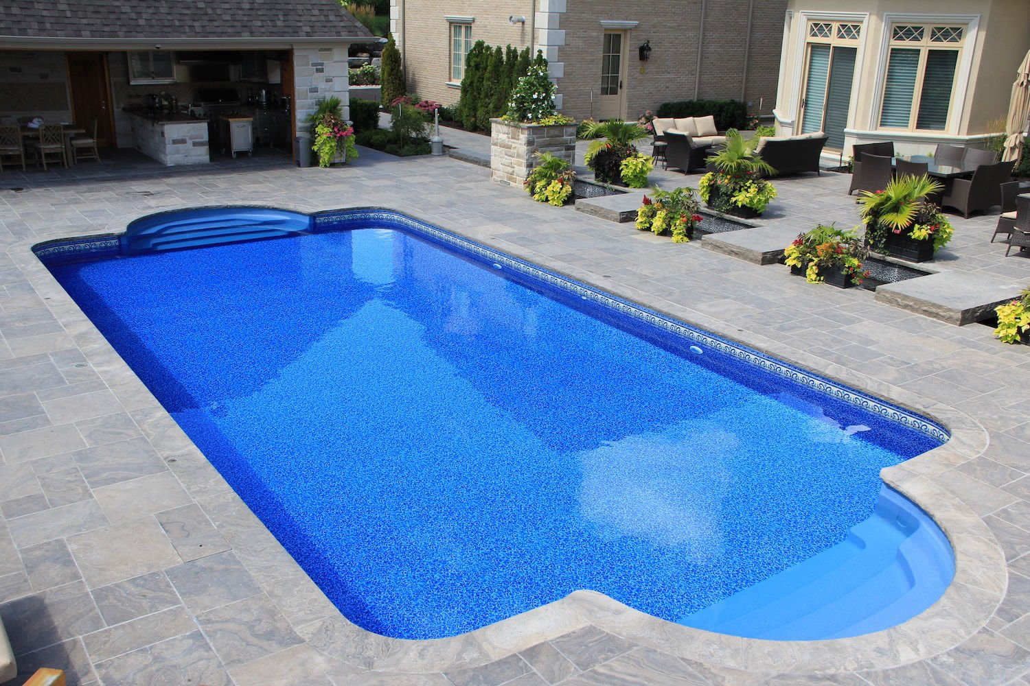 High-angle daytime view of a Roman-style vinyl liner pool featuring vibrant blue water, dual curved end steps, decorative border tile trim, and an expansive stone paver patio.