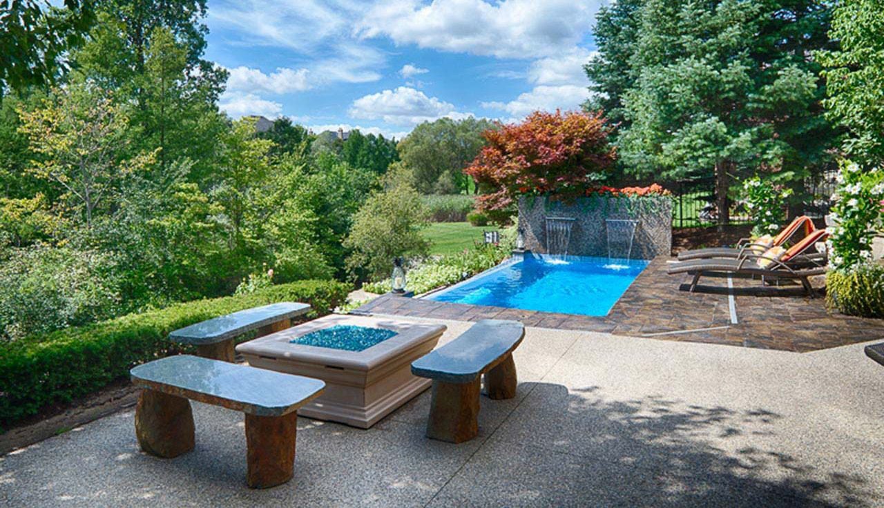 Outdoor installation of a Sea Turtle fiberglass pool featuring dual sheer descent waterfalls, dark paver coping, a fire pit area with stone benches, lounge chairs, and lush surrounding trees.
