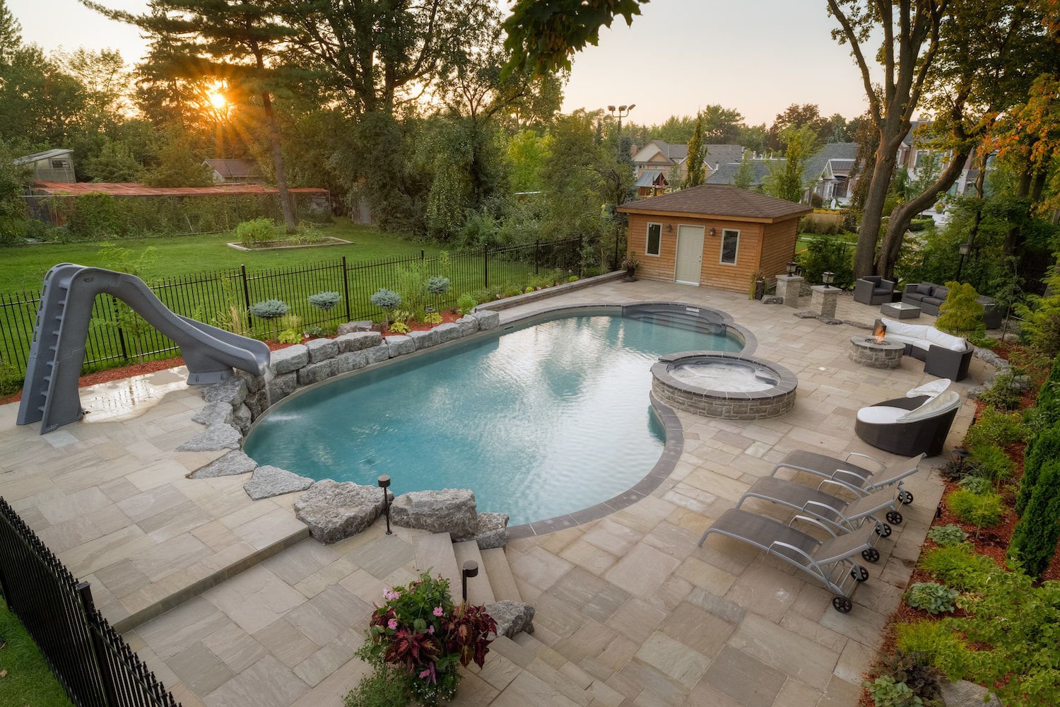 High-angle daytime view of a freeform vinyl liner pool with an integrated raised circular hot tub, a tall water slide built alongside a natural stone retaining wall, stone paver patio decking, and a wooden pool house.