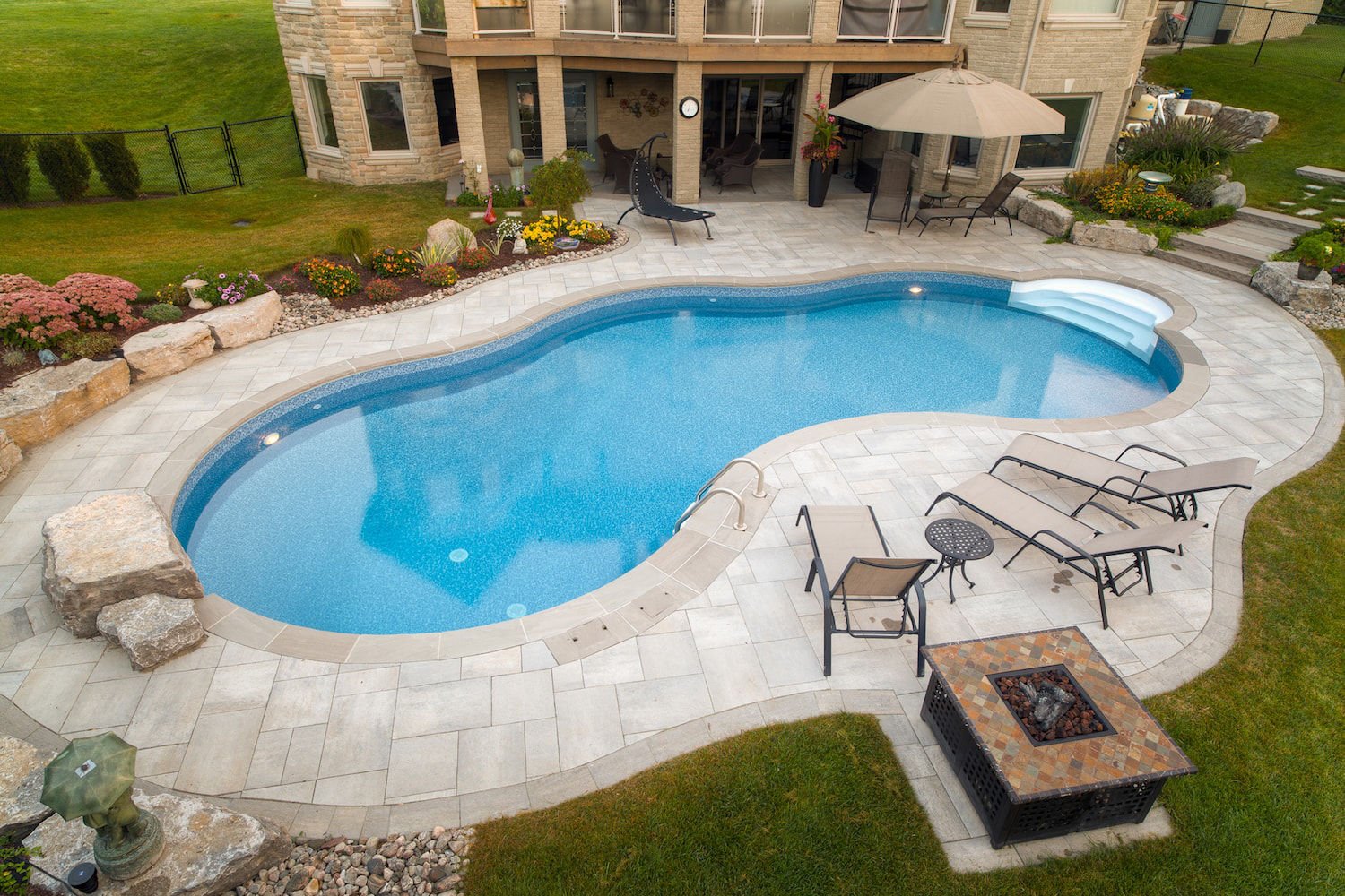 High-angle daytime view of a freeform vinyl liner pool with bright blue water, built-in white acrylic entry steps, smooth stone coping, surrounding light paver patio, poolside lounge chairs, and landscaping.