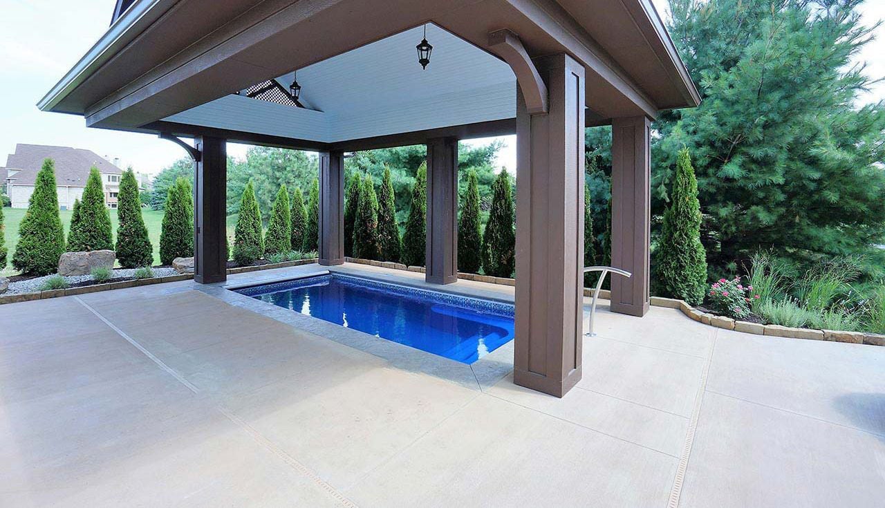 Outdoor view of a rectangular Sea Turtle fiberglass pool sheltered underneath a large brown wooden pavilion structure with pendant lighting, surrounded by concrete decking and privacy evergreen trees.