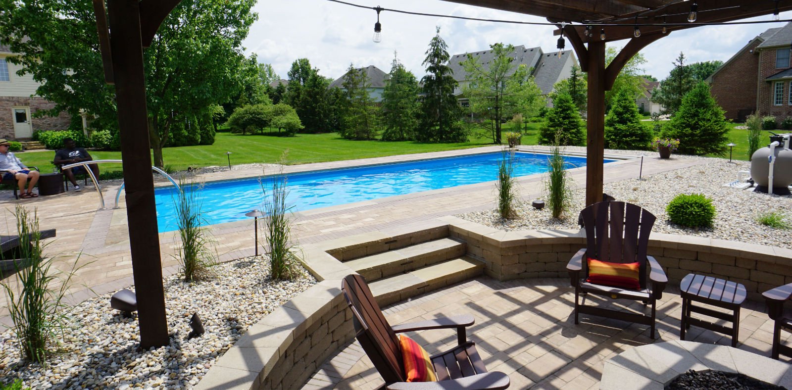 A backyard with a blue swimming pool, surrounded by greenery, outdoor seating, and stone pathways.