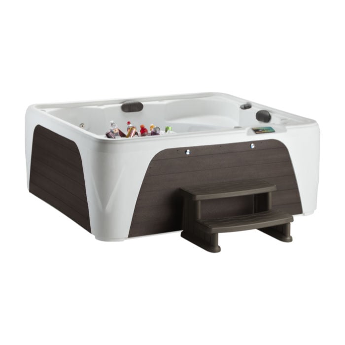 Studio shot of a white Fantasy Spas Enamor Premier hot tub featuring dark wood-grain side panels, matching dark two-tier steps, and a built-in ice bucket filled with drinks.