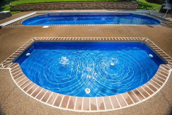 High-angle view of a vibrant blue, semi-circular spa with two bubbling water jets, set into an aggregate patio deck in front of a long rectangular swimming pool.