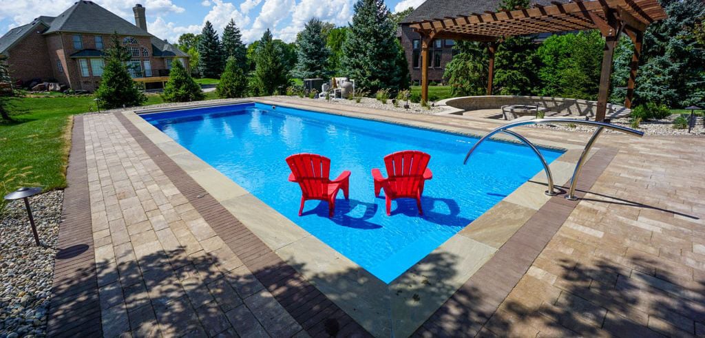 Outdoor backyard pool setting featuring a rectangular bright-blue fiberglass swimming pool with two red Adirondack chairs placed on a built-in tanning ledge, stainless steel handrails, and surrounding stone paver patio.