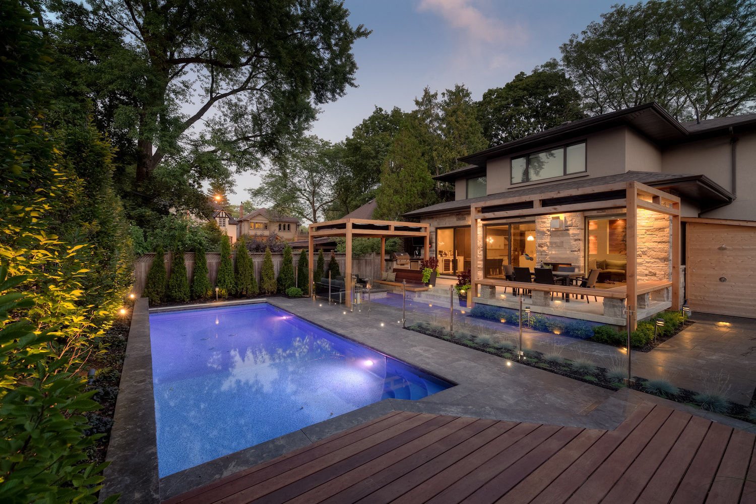 Dusk view of a sleek, rectangular vinyl liner pool illuminated with purple underwater LED lights, featuring dark stone coping, a wooden deck, stone patio, and subtle garden lighting.