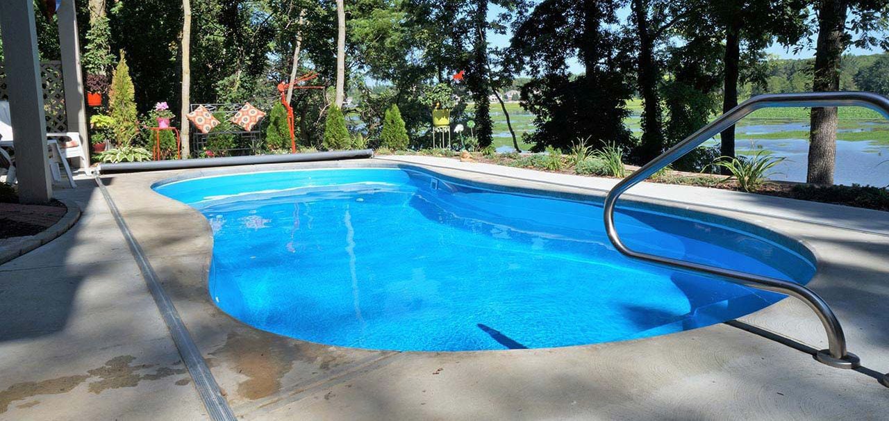 Eye-level view of a curved blue fiberglass pool featuring entry steps, a stainless steel handrail, an automatic pool cover track system, concrete patio decking, and a scenic wooded lake backdrop.