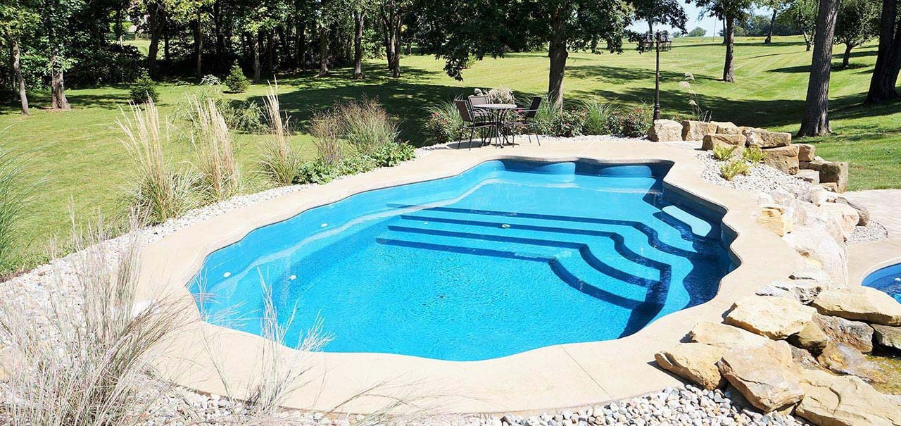 Backyard installation of a scalloped clam shell-shaped fiberglass pool featuring vibrant blue water, concrete coping, tiered entry steps, and surrounding natural rock landscaping.