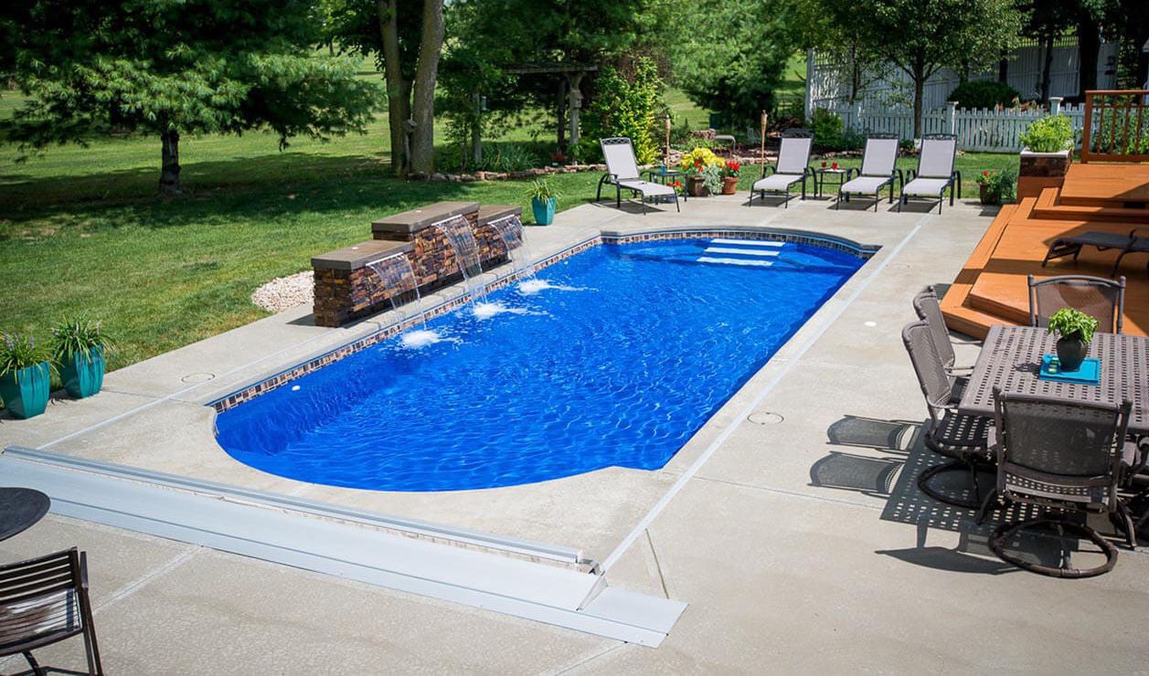 Elevated view of a Cathedral shaped blue fiberglass pool featuring a multi-tiered stacked stone waterfall feature with shear descent cascades, integrated automatic cover, waterline tile, concrete deck, lounge chairs, and patio dining set.