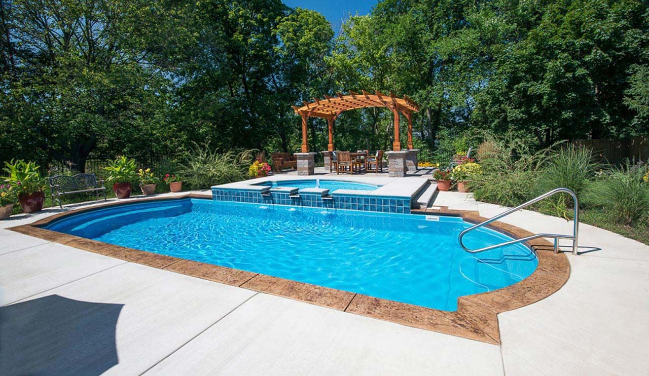 Eye-level view of a classic Cathedral shaped blue fiberglass pool featuring stamped concrete coping, an elevated tiled spa with water spillway features, entry handrail, concrete patio, and a background wooden pergola dining area.
