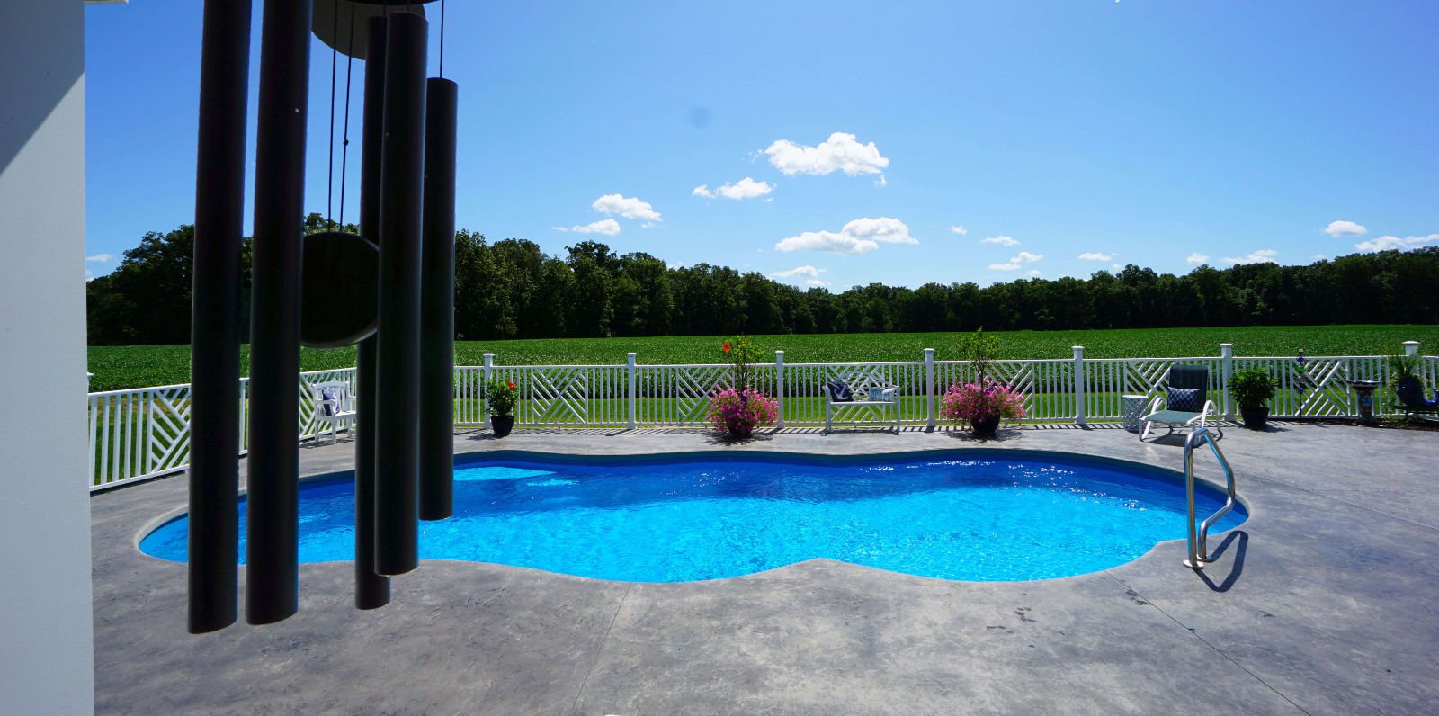 Daytime view of a Thursday Pools Sun Day freeform fiberglass pool featuring vibrant blue water, dark grey stamped concrete patio, white vinyl fencing, wind chimes in the foreground, and scenic open field views.