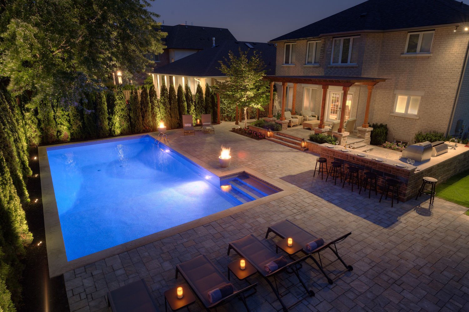 Wide evening shot of a rectangular vinyl liner pool illuminated with blue pool lights, featuring fire bowls, poolside lounge chairs, built-in steps, and surrounding landscape lighting.