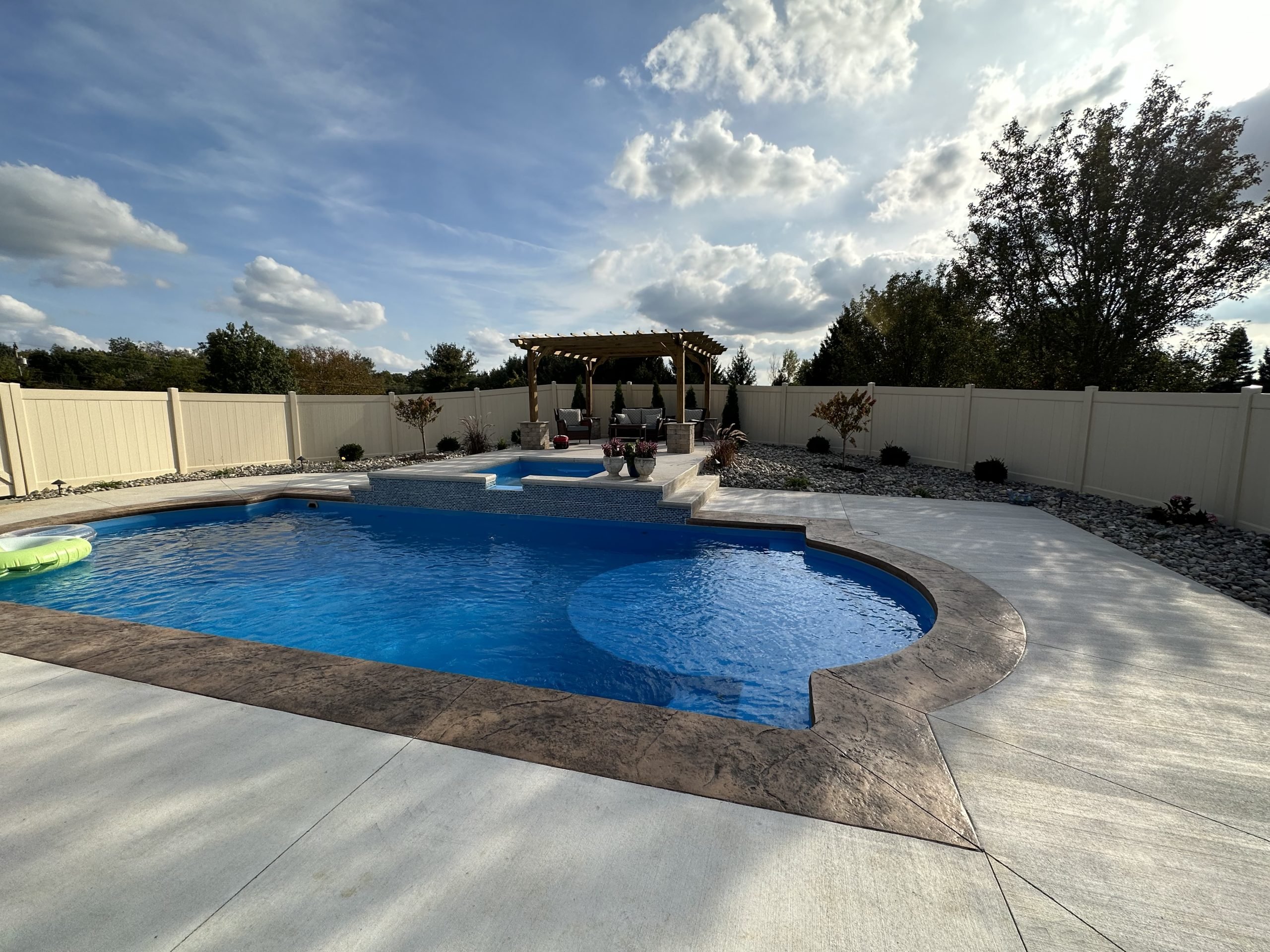 Wide-angle view of a large residential backyard inground swimming pool with dark brown stamped concrete coping, a sun ledge, an elevated spa, and a wooden pergola lounge area inside a fenced yard.