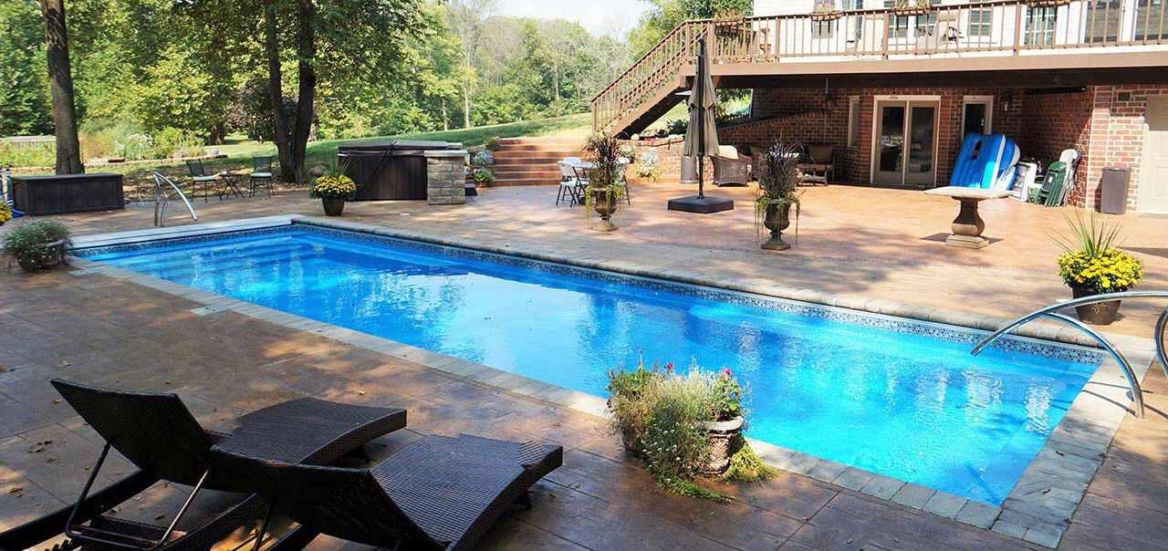 Wide side perspective of a rectangular blue fiberglass pool with full-width entry steps at both ends, stainless steel handrails, decorative waterline tile, and an expansive stamped concrete patio deck adjacent to a walk-out basement.