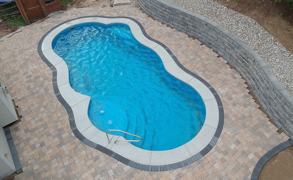 High-angle photograph of a Thursday Pools Sun Day freeform fiberglass pool featuring vibrant blue water, light-colored coping with a dark paver border, paver patio decking, and a curved retaining wall.