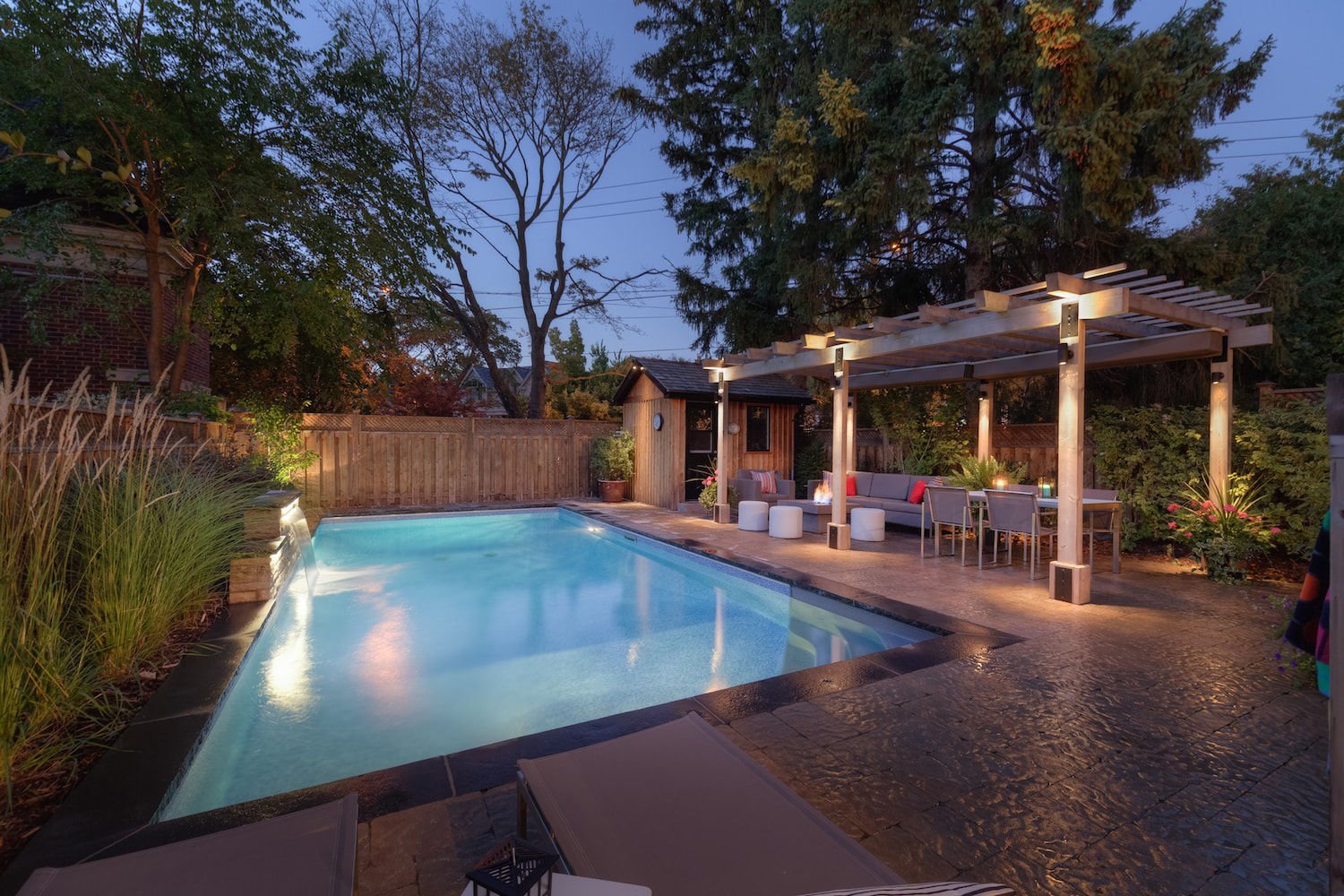 Evening view of a rectangular vinyl liner pool illuminated with soft lighting, featuring a raised stone waterfall feature, dark coping, stamped concrete patio, cabana, and outdoor lounge area.