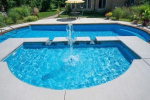 High-angle view of a bright blue semi-circular spa featuring a central water fountain jet, situated on a light concrete patio in front of a matching swimming pool and a lush green backyard.