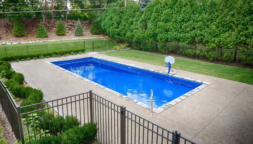 A rectangular swimming pool surrounded by a fence and landscaped greenery, featuring a basketball hoop nearby.