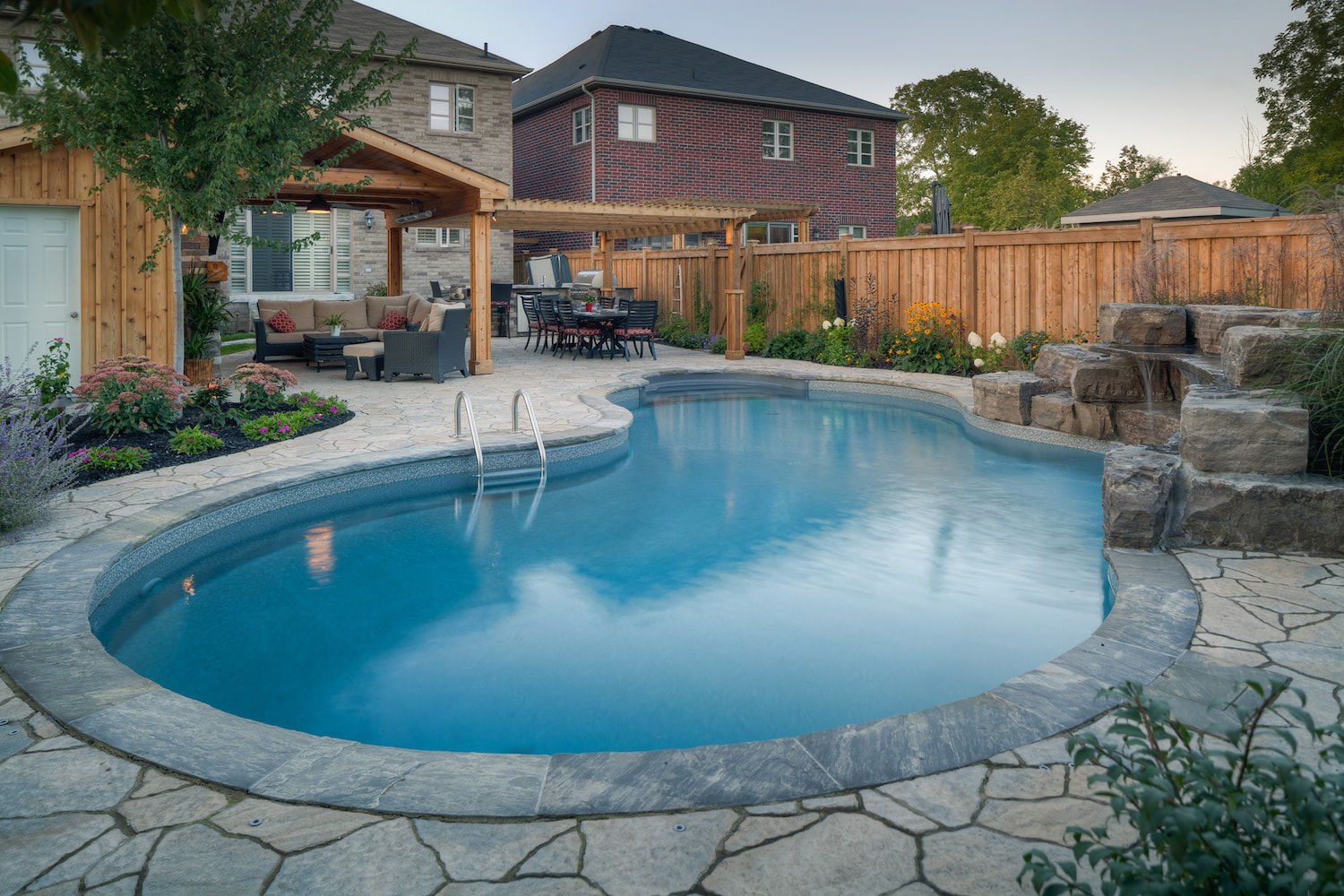 Daytime view of a freeform vinyl liner pool featuring dark stone coping, flagstone patio decking, a natural tiered boulder waterfall, and an outdoor living area with a pergola and seating.