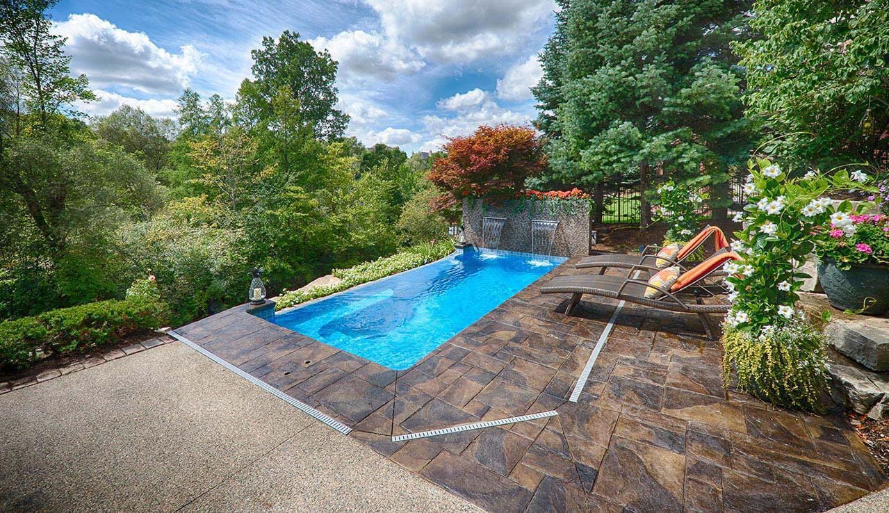 Outdoor view of a rectangular Sea Turtle fiberglass pool featuring dual sheer descent waterfalls, dark stone paver coping, poolside lounge chairs with orange cushions, and lush green forest landscape under a blue sky.