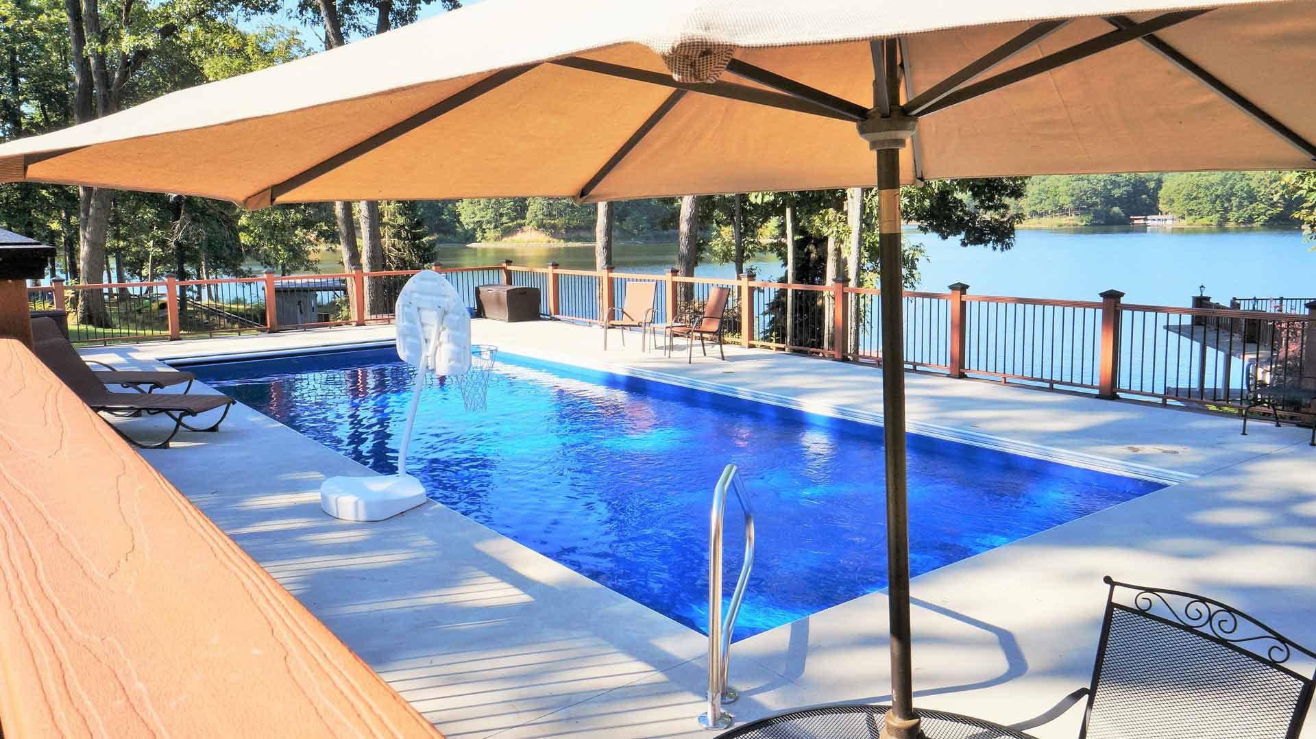 A swimming pool with a blue surface, surrounded by trees and a lake view, under a shaded patio umbrella.