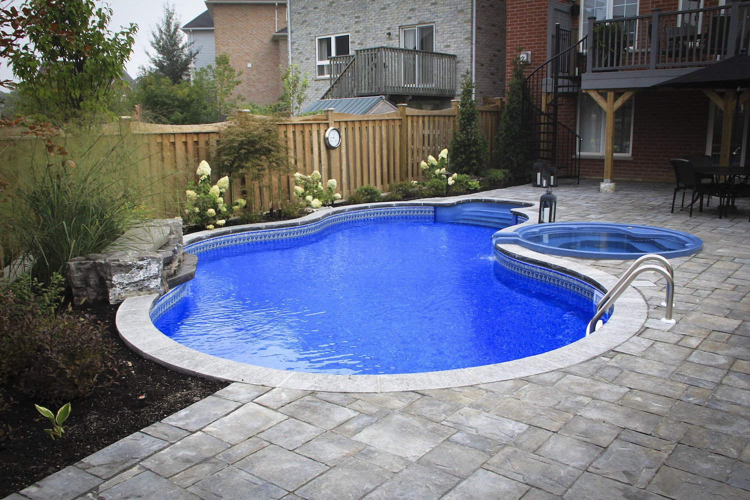 Daytime view of a freeform vinyl liner pool featuring vibrant blue patterned water, light bullnose coping, an integrated circular spillover hot tub, built-in alcove steps, a small natural rock waterfall, and stone paver decking.