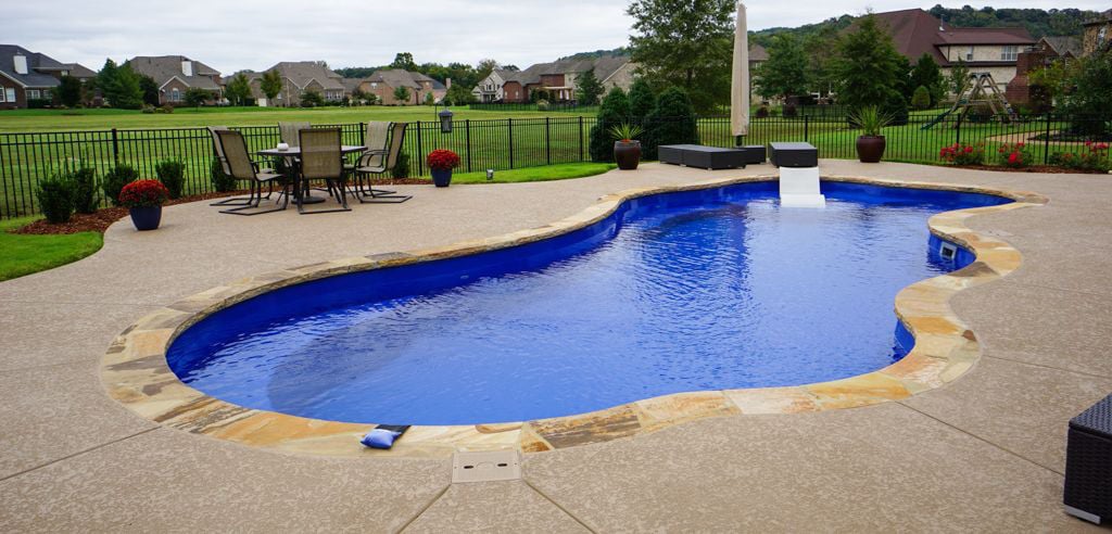 Eye-level view of a freeform blue fiberglass pool featuring natural flagstone stone coping, an integrated tanning ledge with a lounger, textured concrete patio decking, outdoor dining table, and wrought-iron fencing.