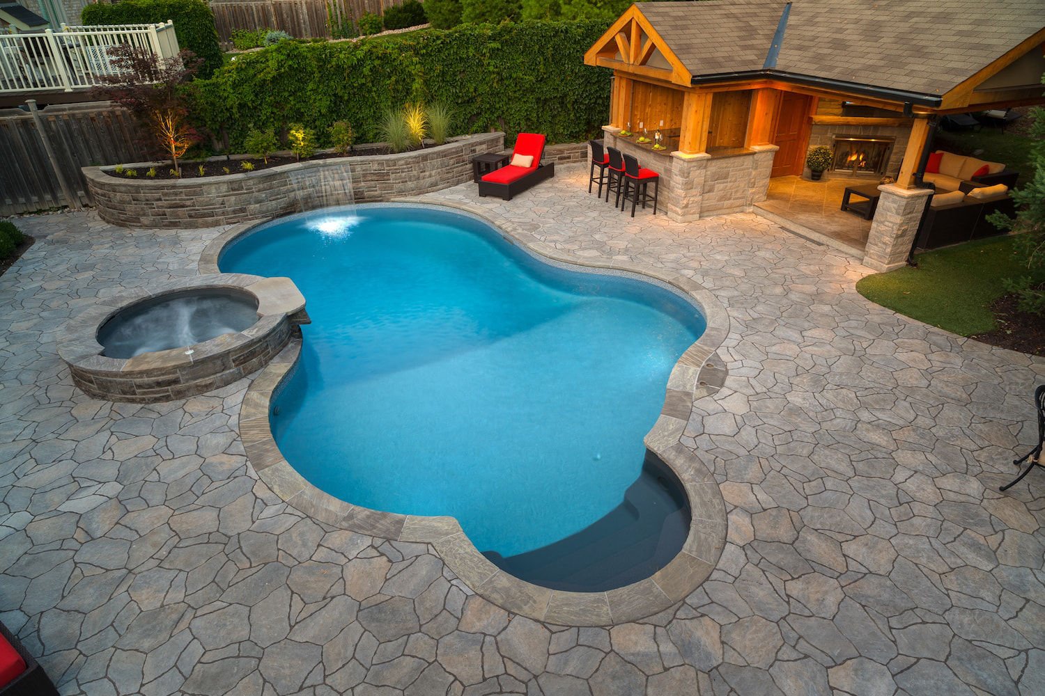 High-angle view of a freeform vinyl liner pool featuring a raised circular stone hot tub, sheer curtain waterfall from a retaining wall, dark entry steps, flagstone patio decking, and outdoor bar seating.