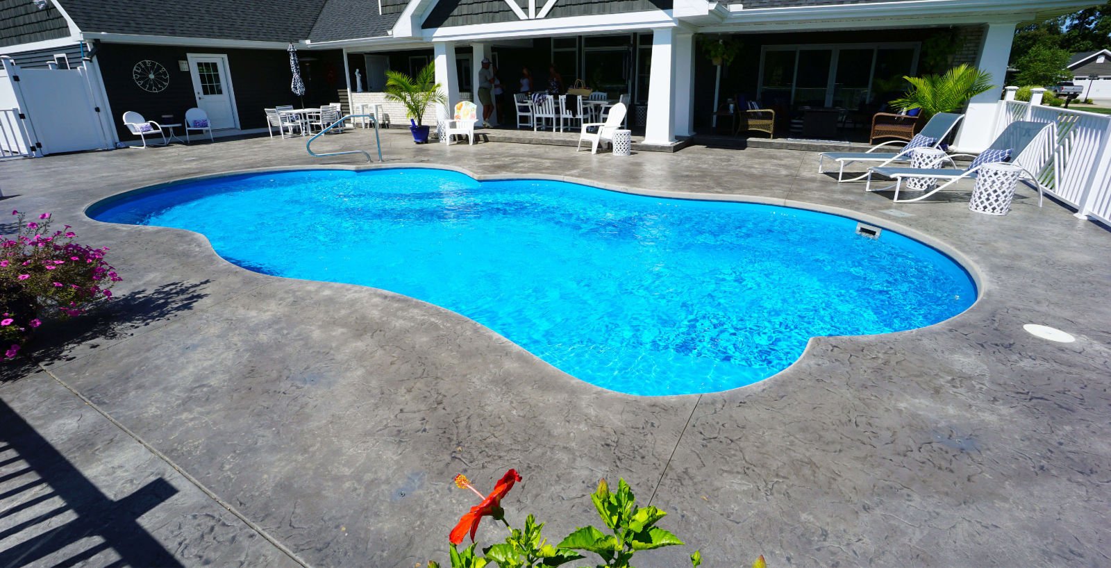 Outdoor installation of a Thursday Pools Sun Day freeform fiberglass pool featuring clear blue water, grey stamped concrete decking, surrounding lounge chairs, and a white residential home backdrop.