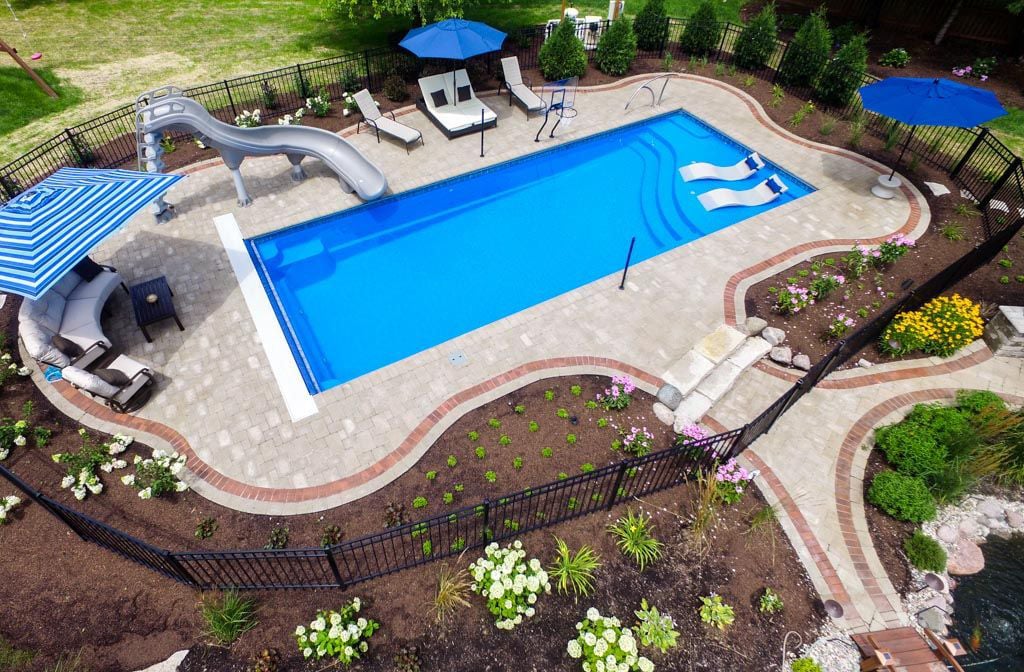 Aerial view of a backyard pool with a slide, surrounded by lounge chairs, umbrellas, and flower beds.